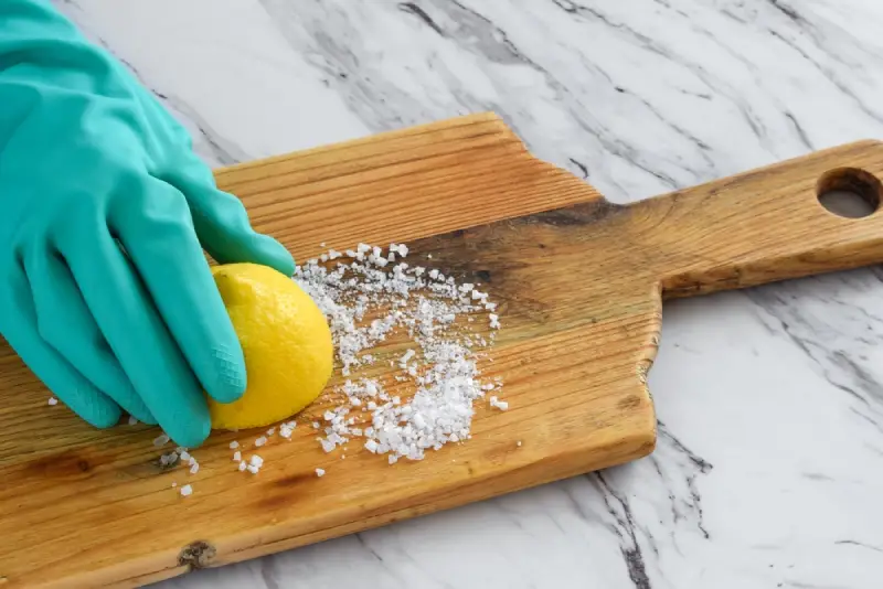 Closeup of person&rsquo;s hand in protective rubber glove scrubbing wooden cutting board with lemon juice and salt